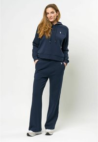 Tumši zilas krāsas kapuce un saskaņoti bikses, izgatavotas no mīksta auduma, ar kengura kabatu, regulējamu kapuci un izšūtu logo detaļu.