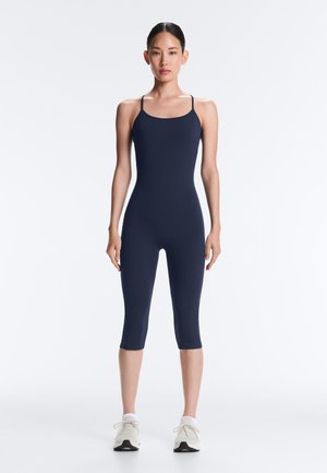 Žena stojaca tvárou dopredu, oblečená v tmavomodrý priliehavý overal bez rukávov a biele tenisky na jednoliatom bielom pozadí.
