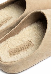 Zapatillas de ante beige con un suave forro de piel de oveja color crema. Presentan un diseño moldeado con una pequeña etiqueta de marca cosida.