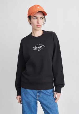 Άτομο με πορτοκαλί καπέλο, μαύρο φούτερ Carhartt και μπλε τζιν, κοιτάζοντας προς το πλάι με φόντο έναν απλό φωτεινό τοίχο.