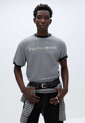 Fiatal férfi csíkos "FULFILLMENT" feliratú pólóban, fekete farmerben, övvel és kockás inget a derekán megkötve, egyszerű háttér előtt állva.