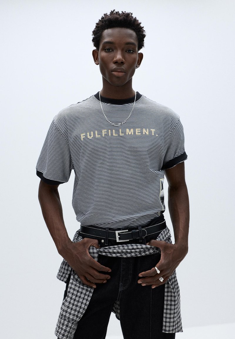 Jeune homme portant un t-shirt rayé avec l'inscription "FULFILLMENT", un jean noir, une ceinture, et une chemise à carreaux nouée autour de la taille, debout devant un fond uni.