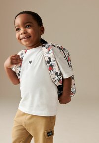 Sac à dos avec un motif coloré de Mickey Mouse, avec des bretelles réglables, associé à un t-shirt blanc uni et un pantalon beige.