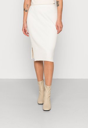 Kvinde iført en hvid ribbet midi-nederdel med sideslidser, beige ankelstøvler og synlige tatoveringer på armene.