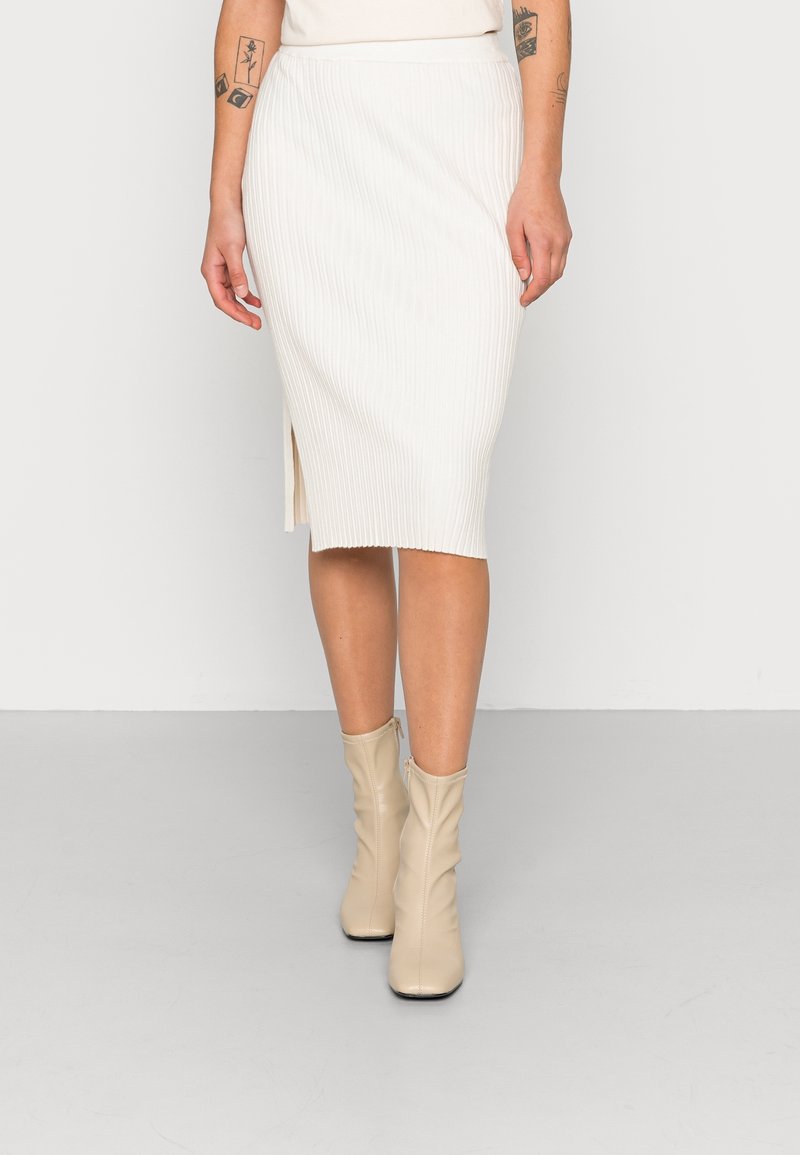 Femme portant une jupe midi blanche côtelée avec une fente latérale, des bottines beiges et des tatouages visibles sur les bras.