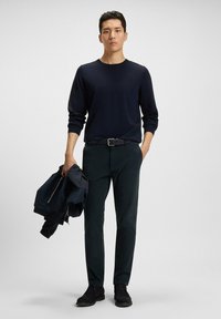 Navy crew-neck sweater, dark green slim-fit trousers, black belt, and ankle boots; holding a navy bomber jacket. Smooth texture, minimalist design.
