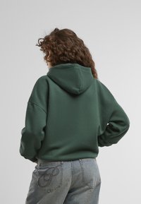 Mørkegrøn hætte sweatshirt med en stor hætte og sænkede skuldre, parret med lyseblå jeans, der har en broderet detalje på baglommen.