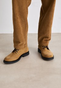Des chaussures en daim tan avec des semelles en caoutchouc noir, dotées de lacets et d'un bout arrondi, associées à un pantalon en velours côtelé marron à nervures.
