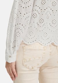 Weiße Bluse mit Lochstickerei, gewelltem Saum, langen Ärmeln und Knopfmanschetten, kombiniert mit hellbeigen Jeans, die über eine Gesäßtasche verfügen.