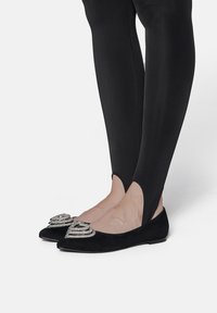 Ballerines en cuir suédé noir avec des pointes effilées et des accents décoratifs métalliques argentés à l'avant. Portées avec des leggings noirs ajustés.