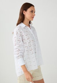 White button-up shirt with eyelet design, long sleeves, and a collar. The fabric has a floral pattern of perforations with subtle texture.