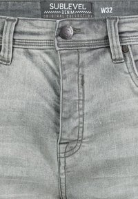 Gray denim jeans with a faded finish, featuring a front button closure and five-pocket design. Label reads "SUBLEVEL W32."