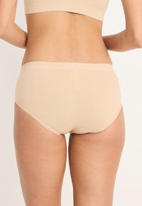 Vrouw in beige naadloze ondergoed en bijpassende beha, gezien vanaf de achterkant tegen een effen lichte achtergrond.