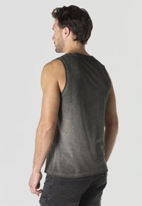 Hombre vistiendo una camiseta sin mangas negra descolorida y vaqueros oscuros, visto desde atrás sobre un fondo liso.