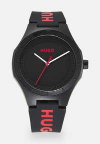 Black rubber watch with a round face, red accent detailing, white hour and minute hands, and bold "HUGO" text on the strap.