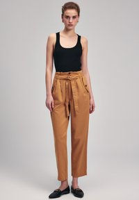Zwarte geribbelde tanktop, hooggezette mosterdkleurige broek met een gestrikt riem, zijzakken en zwarte instappers tegen een neutrale achtergrond.