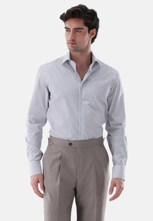 Mann trägt weißes Langarmhemd mit dünnen schwarzen vertikalen Streifen und beigefarbene, hoch taillierte Anzughose, schaut zur Seite.