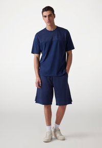 Marine atletikoutfit bestående af en kortærmet, struktureret skjorte og knælange shorts, begge lavet af åndbart materiale. Hvide sneakers bæres.