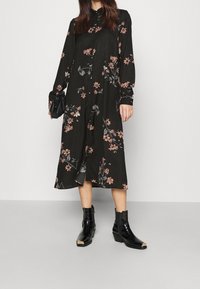 Nainen, joka käyttää mustaa kukallista midi-mekkoa pitkillä hihoilla, pitää mustaa clutch-laukkua ja käyttää mustia nilkkureita metallisten kärjillä.