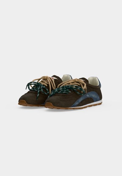 Paire de baskets en daim marron foncé avec des accents bleus, des lacets bicolores beige et verts, et des semelles blanches sur fond blanc.