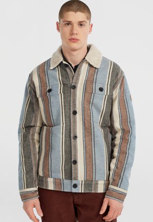 Jeune homme portant une veste à rayures avec un col en fourrure et un pantalon marron, se tenant devant un fond blanc.