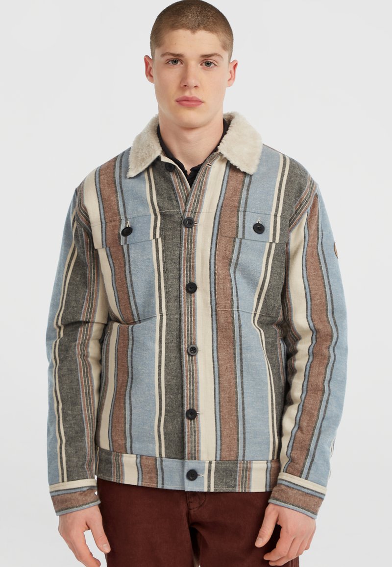 Jeune homme portant une veste à rayures avec un col en fourrure et un pantalon marron, se tenant devant un fond blanc.