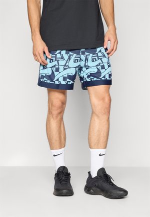 Blauwe en marineblauwe zwembroek met geometrische patronen, gecombineerd met zwarte sportschoenen en witte sokken met een logo.