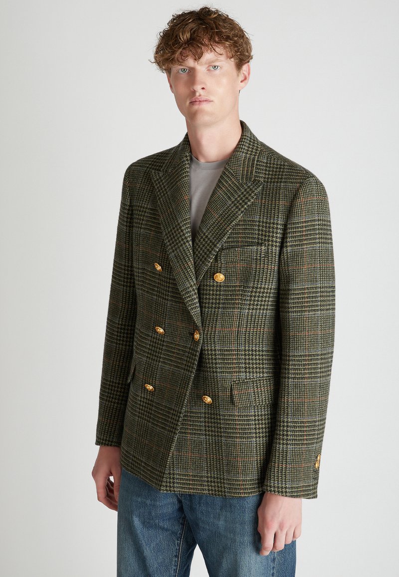 Blazer doppiopetto in plaid verde e marrone, realizzato in lana texture con dettagli di bottoni dorati, abbinato a una camicia grigia e jeans.