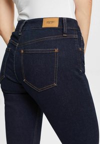 Zblízka člověk v tmavě modrých džínech se oranžovým prošíváním a hnědým nášivkem s logem Esprit New York na pase.