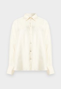 Chemise à manches longues de couleur crème, boutonnée, avec col pointu et poignets boutonnés, présentée sur un fond gris clair uni.