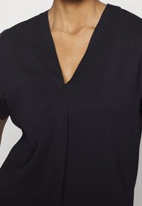 Vêtement noir à encolure en V avec manches courtes, présentant une texture lisse et un design minimaliste. Le tissu semble léger et respirant.