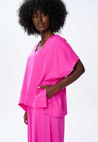 Różowa, luźna bluzka z dekoltem w serek i krótkimi rękawami, z asymetrycznym dołem i kieszonką. Jedwabista tekstura, noszona na pasujących spodniach.