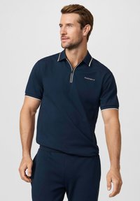 Námořnická polokošile se zipovým límcem a bílými akcenty. Na levé straně hrudi je bílý nápis "Hackett". Materiál působí hladce a lehce.