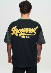 T-shirt noir oversize avec des manches vertes et grises. Présente un texte jaune "Recovered" et des logos de football au dos. Matériau en coton doux.