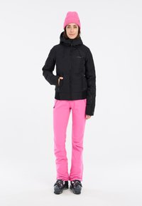 Schwarz isolierte Jacke mit Kapuze, kombiniert mit leuchtend rosa Skihosen. Das Outfit umfasst schwarze Skischuhe mit einem gemusterten Design.