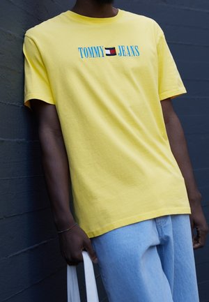 Mies pukeutuneena keltaiseen Tommy Jeans -t-paitaan ja vaalean sinisiin farkkuihin, pitää valkoista kangaskassia, seisoo tummanharmaan seinän edessä.