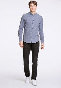 Niebieska koszula zapinana na guziki z fakturowanej tkaniny, długimi rękawami i czarnymi spodniami typu slim-fit. Stylizowana z białymi sneakersami, stojąca na jednolitym tle.