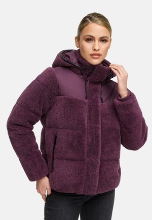Femeie atingând gulerul unei jachete din fleece violet cu glugă, stând pe un fundal simplu de culoare deschisă.