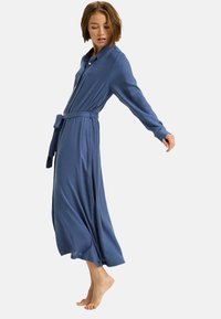 Langes blaues Kleid aus fließendem Stoff, mit einem geknöpften Kragen, langen Ärmeln und einer taillierten Taille. Gelegentliche seitliche Schlitze sorgen für Bewegung.