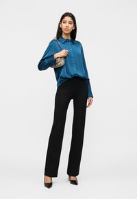 Γυναίκα που στέκεται φορώντας μια σατέν μπλούζα σε τυρκουάζ, μαύρο παντελόνι, μαύρες γόβες και κρατώντας μια μικρή μπεζ τσάντα ώμου.