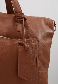 Bolso tote de cuero marrón con superficie texturizada, un bolsillo con cremallera y una etiqueta de equipaje con detalles de flecos.
