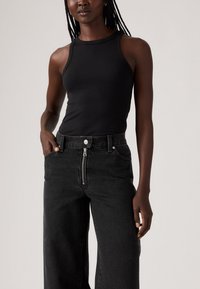 Top in cotone nero a costine abbinato a jeans neri a vita alta con zip e tasche; il modello posa con una mano sui fianchi.