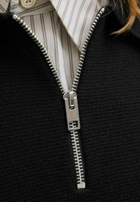 Detailní záběr na stříbrný zip na černém pleteném svetru, který je navlečený přes bílou košili s pruhy.