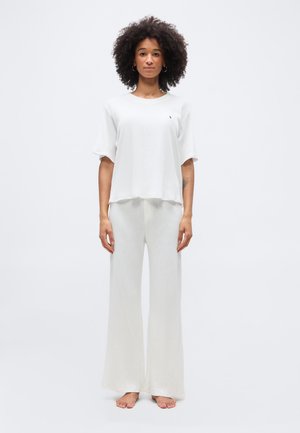 Femeie cu păr creț stă desculță, purtând un top alb larg cu mâneci scurte și pantaloni albi evazați, pe un fundal simplu și deschis la culoare.