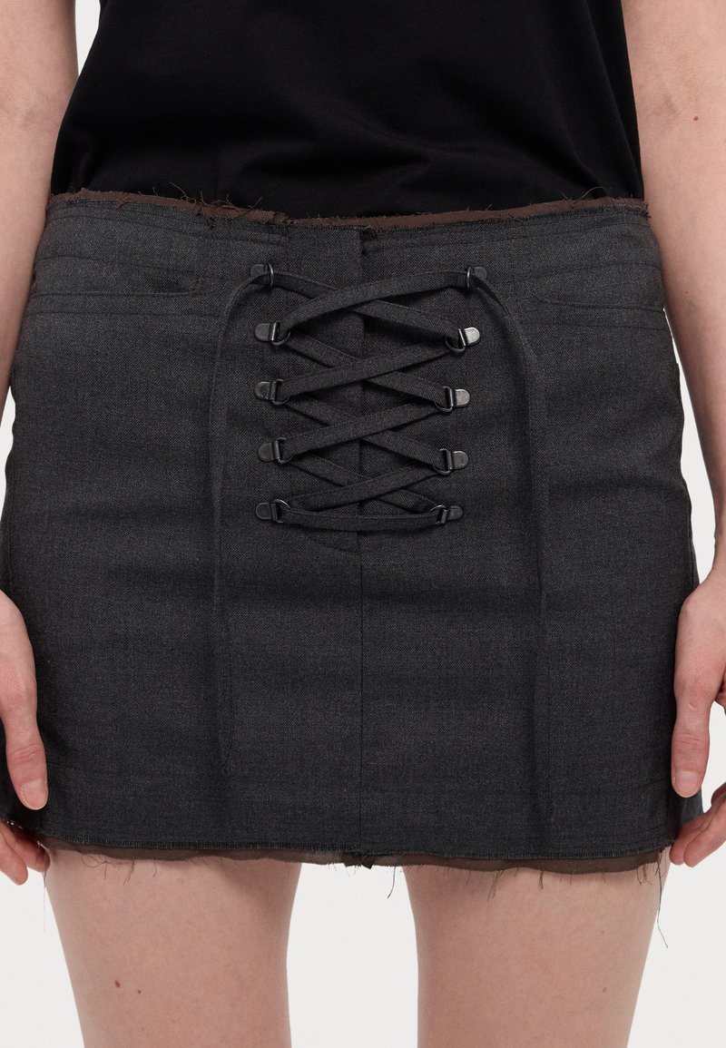 Gonna mini grigia scura realizzata in materiale texturizzato, con un design anteriore con lacci e hardware nero, e dettagli di orlo sfrangiato.