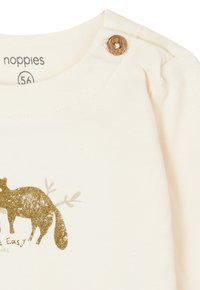 Helles beige Baumwollhemd mit einem grafischen Aufdruck eines goldenen und grauen Fuchses, ergänzt durch Blätter; verfügt über einen Holzknopf nahe der Schulter zur Befestigung.