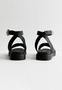 Sandalias de cuero negro con un acabado texturizado, que presentan tiras entrecruzadas y un acento de hebilla dorada en la correa del tobillo. Diseño de suela plana.