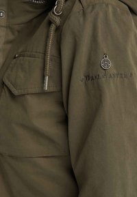 Manica di giacca verde oliva con il testo ricamato "Drill Master", charm metallico rotondo con ancora e dettagli con cordino intrecciato con punte in metallo.