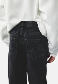 Sorte denim jeans med høj talje, der har to baglommer med tonet broderi. Bæres med en hvid sweatshirt.