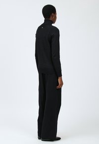 Schwarzer Strickrollkragenpullover mit geripptem Saum und Ärmeln, einem subtilen metallischen Akzent auf der Rückseite, kombiniert mit weit geschnittenen schwarzen Hosen.